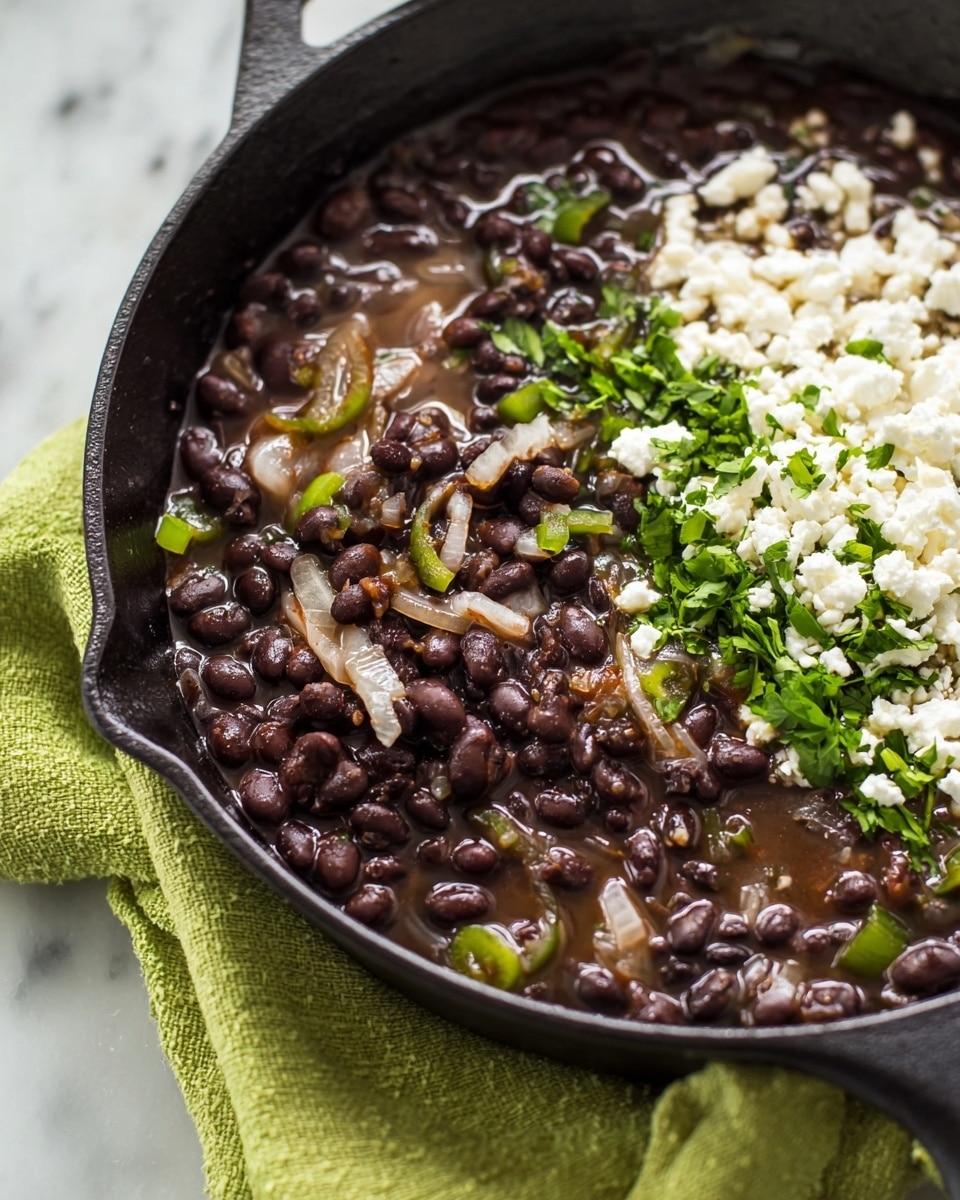 The image shows a close-up of a black cast iron skillet filled with black beans cooked in a brown broth with translucent cooked onion strips and small green vegetable pieces. A small amount of crumbled white cheese and chopped green herbs are on one side of the beans, adding texture and color contrast to the dish. The skillet rests on a pale green textured cloth, all set against a white marbled surface. photo taken with an iphone --ar 4:5 --v 7