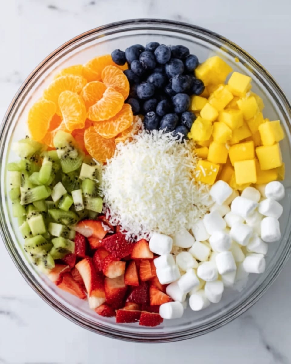 A clear glass bowl filled with a colorful fruit and marshmallow salad sits on a white marbled surface. The bowl is divided into neat sections: bright orange mandarin slices on the top left, small dark blue blueberries next to them, and finely chopped green kiwi pieces in the top right. Two sections of yellow mango chunks are placed next to the kiwi on the right and on the left side below the blueberries. Red strawberry pieces form the bottom left section, and small white marshmallows fill the bottom right section. In the center, a small pile of shredded white coconut adds texture and contrast to the colorful layers around it. Photo taken with an iphone --ar 4:5 --v 7