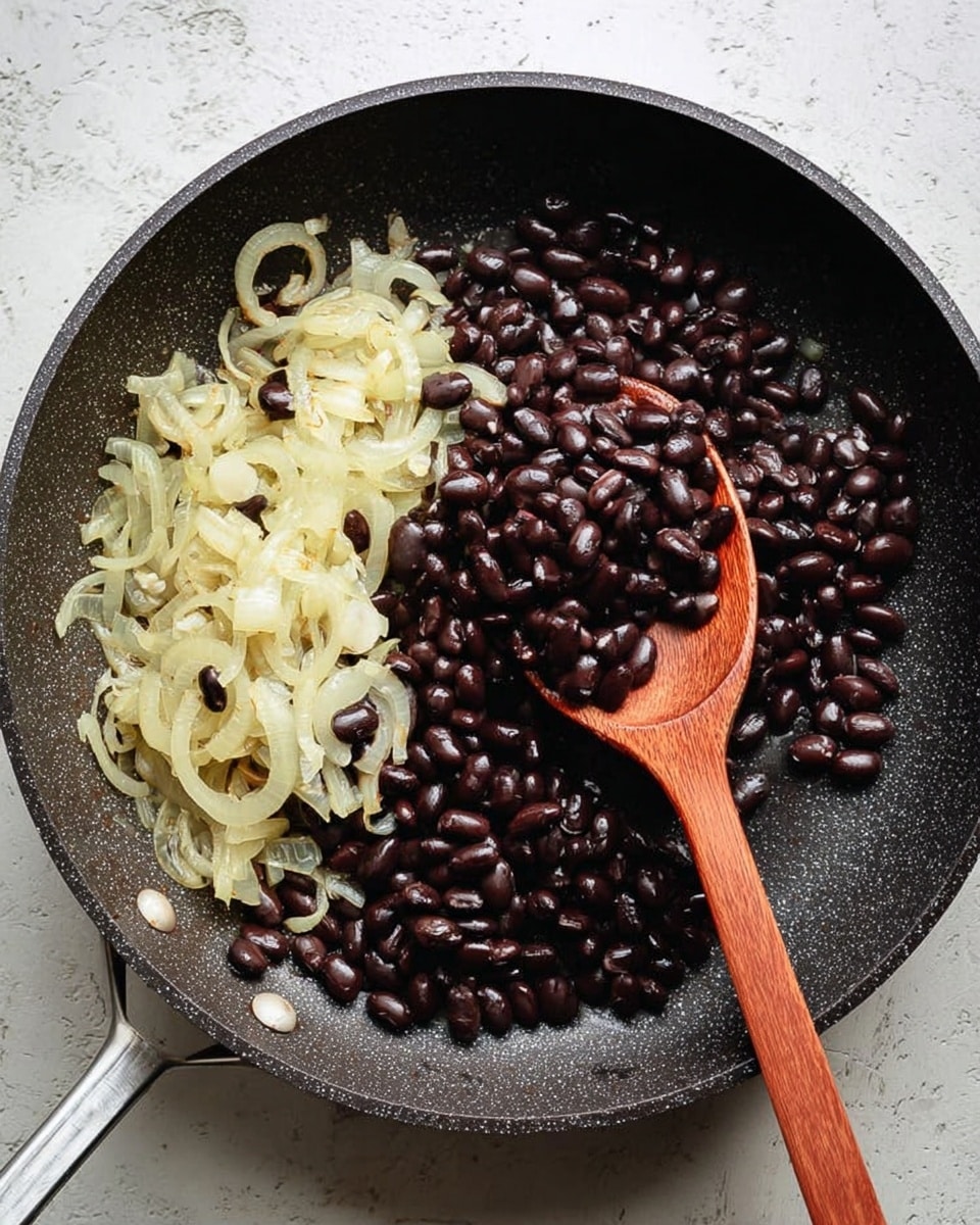 A black frying pan holds two main layers from left to right: tender, light golden sautéed onions with a soft texture, and shiny, dark black beans with a smooth surface. A wooden spoon rests on the onions, slightly lifting some of them. The pan sits on a white marbled textured surface. Photo taken with an iphone --ar 4:5 --v 7