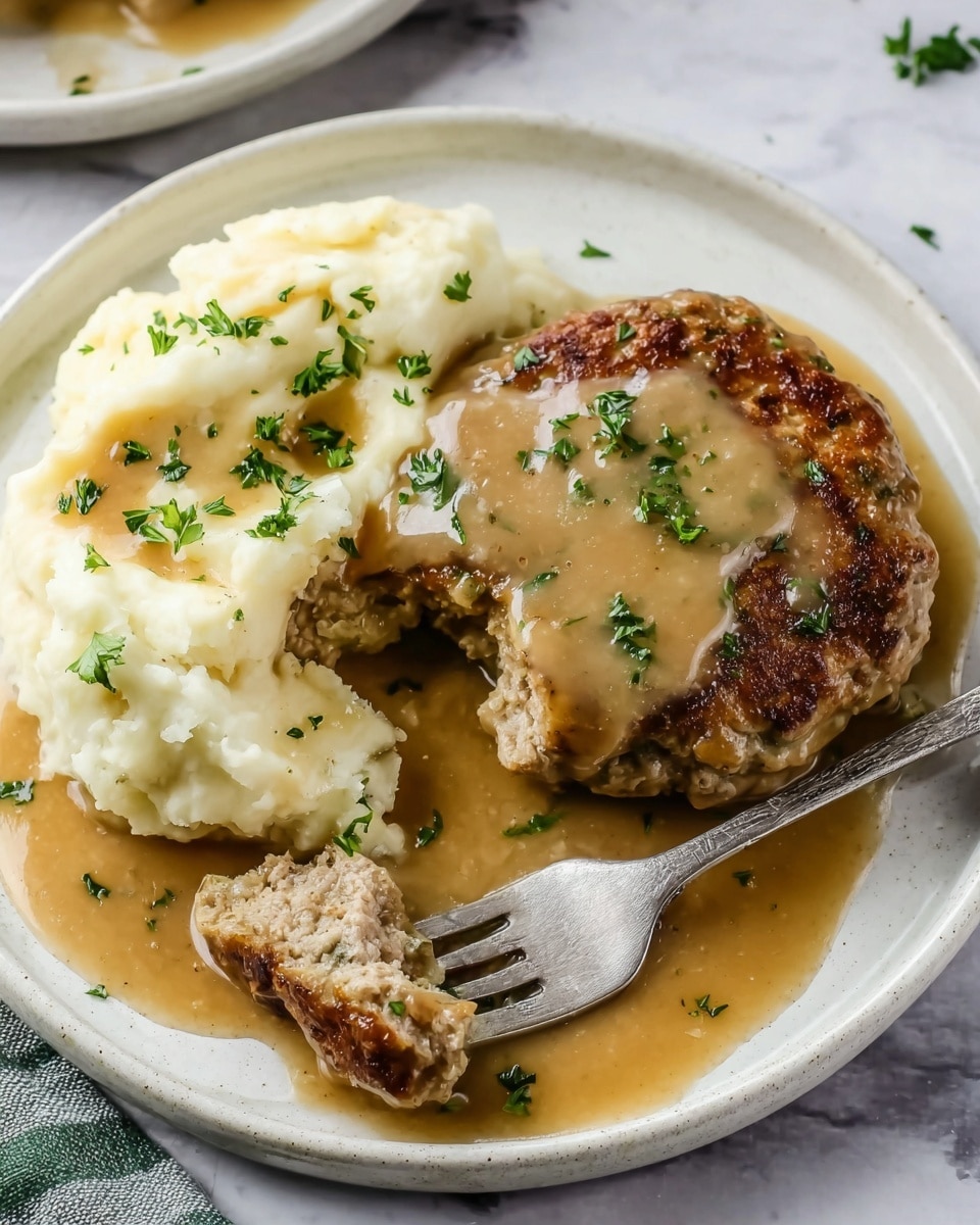 A white plate holds a serving of mashed potatoes topped with light brown gravy and sprinkled with green parsley. Next to the potatoes is a round browned meat patty covered in the same gravy and garnished with green parsley. A fork with a piece of the meat patty on it rests on the plate beside the patty. The plate is set on a white marbled surface. Photo taken with an iphone --ar 4:5 --v 7