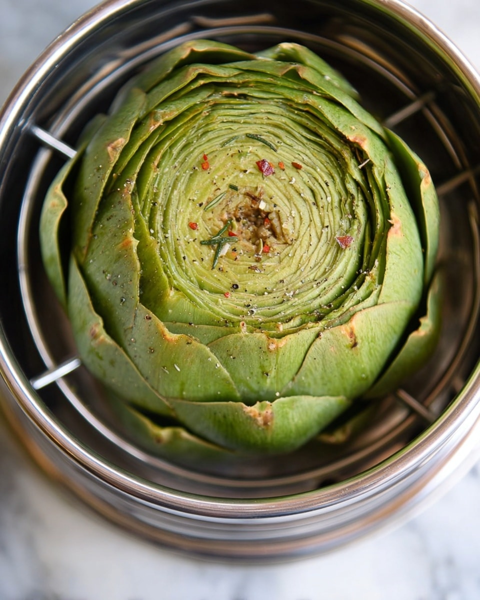 A single large green artichoke sits inside a round metal steamer basket with metal bars at the bottom. The artichoke petals are tightly packed and thick, showing layers of light to dark green shades from the center outwards. The top is sprinkled with small flecks of black pepper, dried herbs, and red spices. The metal steamer has a shiny silver edge and rests on a white marbled surface. Photo taken with an iphone --ar 4:5 --v 7