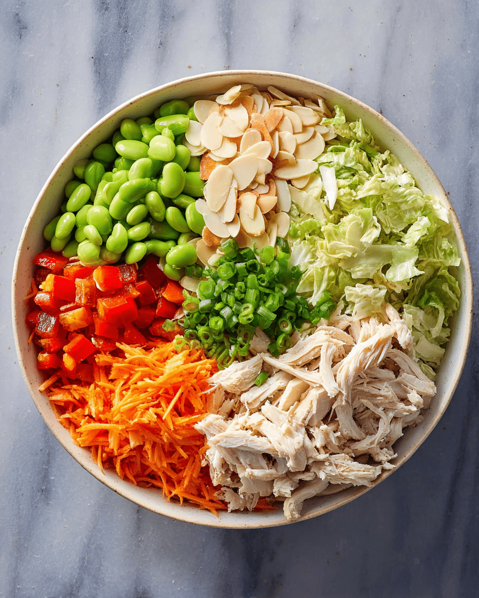 A white bowl holds six colorful layers arranged in sections. Starting from the bottom right, there is a pile of light beige shredded cooked chicken. To the right of the chicken, there is a layer of chopped light green lettuce. Above that, a small section of thin, sliced almonds adds a creamy white texture. Next to the almonds, diced red bell peppers form a bright red block. To the left, green edamame beans cover a medium section. Below the beans is a pile of bright orange shredded carrots. Finally, above the chicken and carrots, chopped green onions rest in the center. The bowl sits on a white marbled surface. Photo taken with an iphone --ar 4:5 --v 7