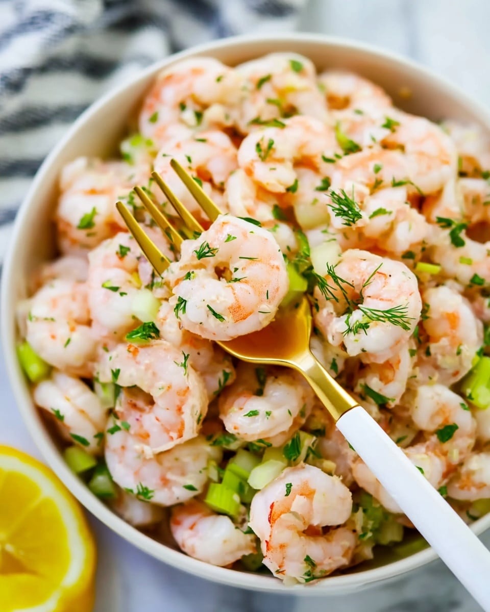 A bowl filled with many cooked shrimp, light pink in color with small green herb leaves sprinkled on top. Beneath the shrimp, there are small white and light green chopped pieces that look like celery and onion. A golden fork with a white handle is picking up some shrimp and celery from the bowl. The bowl is white and placed on a white marbled surface, with a small slice of bright yellow lemon visible in the bottom left corner of the image. photo taken with an iphone --ar 4:5 --v 7