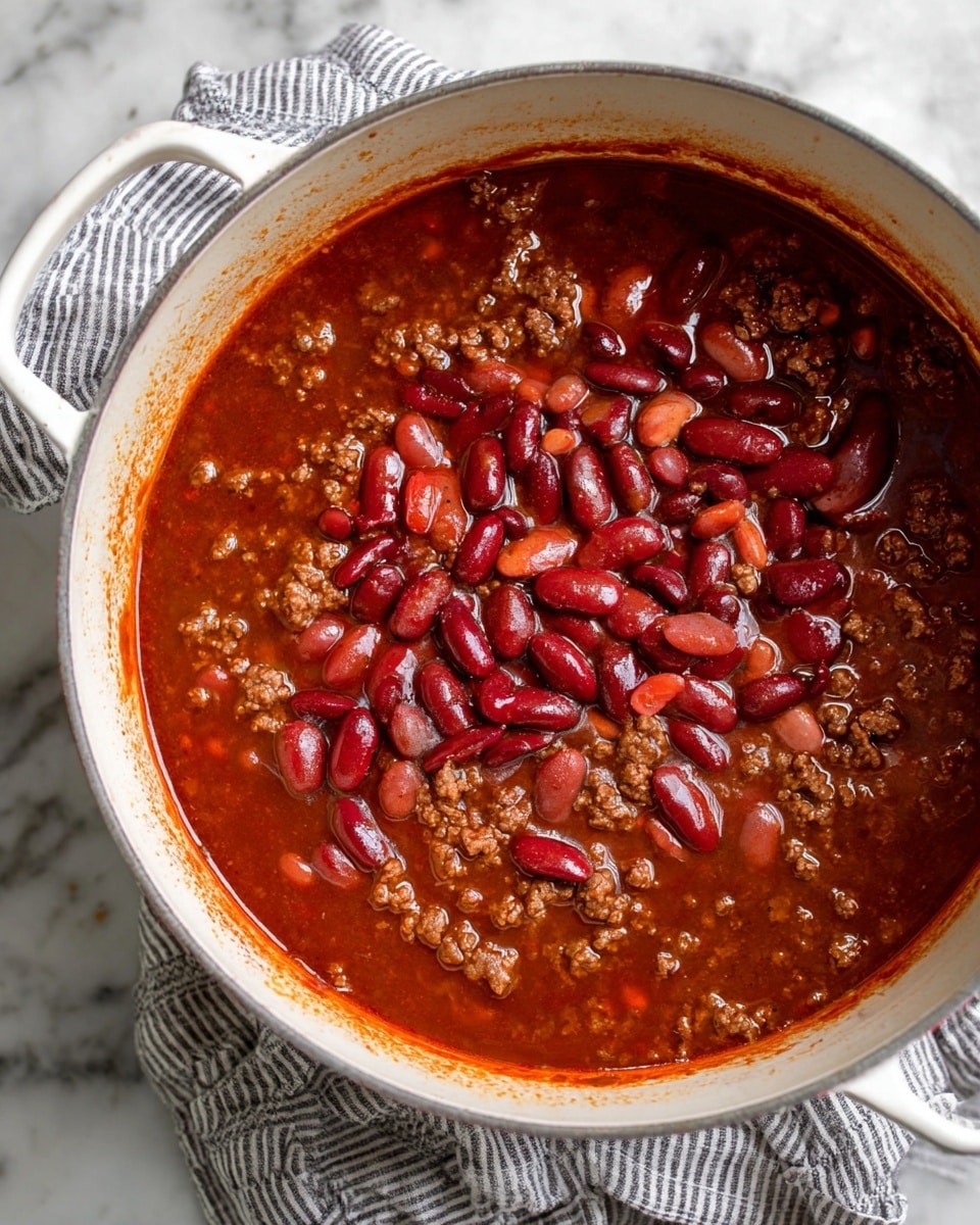 A white pot filled with a thick dark red chili sauce as the base layer covering the entire bottom and sides, mixed with small chunks of ground meat scattered unevenly around the edges. On top, a mix of light and dark red kidney beans sitting in a shallow layer of reddish-brown liquid, some beans floating and some slightly submerged, creating a shiny and wet texture. The pot rests on a striped cloth, placed on a white marbled surface. Photo taken with an iphone --ar 4:5 --v 7