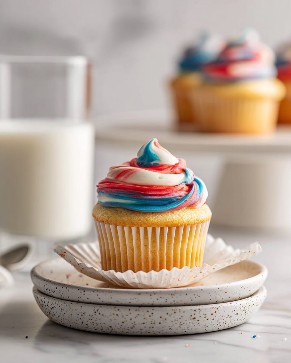 A single vanilla cupcake with three layers of frosting swirled on top in red, white, and blue colors. The cupcake is placed in a white paper liner that is slightly open, sitting on two stacked white speckled plates. In the background, there is a clear glass filled with milk and a blurred white cupcake stand holding another cupcake with the same frosting colors. The whole scene is set on a white marbled surface with a soft, bright light giving a fresh feel. photo taken with an iphone --ar 4:5 --v 7