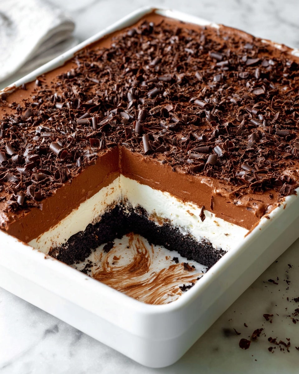 The image shows a multi-layered chocolate dessert in a white rectangular dish, with one large piece removed from the front right corner. The bottom layer is a dark cookie crumb base, followed by a thick white creamy layer. Above that is a smooth, thick milk chocolate layer, topped by another white creamy layer. The top layer is covered with finely crushed dark cookie crumbs and sprinkled with many small chocolate curls, creating a textured, rich dark brown surface. The dish sits on a white marbled surface. Photo taken with an iphone --ar 4:5 --v 7