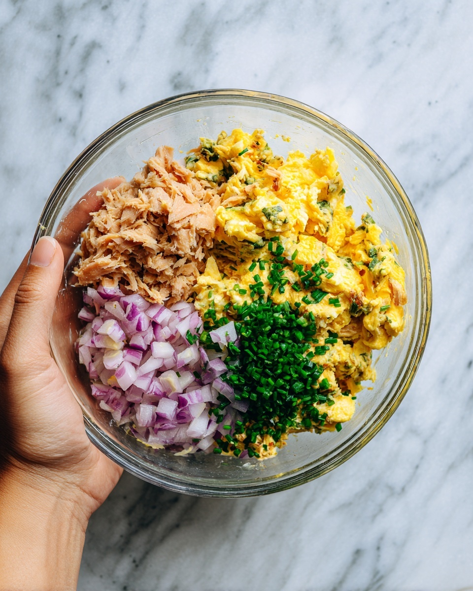A clear glass bowl held by a woman's hand shows four distinct layers of food inside. The top left layer is light brown, flaky tuna. The top right layer is soft, yellow scrambled eggs with a slightly moist texture. The bottom left layer contains small, diced red onion pieces, purple and white in color. In the center is a small pile of finely chopped green herbs, likely chives, adding freshness and color contrast. All this is against a white marbled surface background. photo taken with an iphone --ar 4:5 --v 7