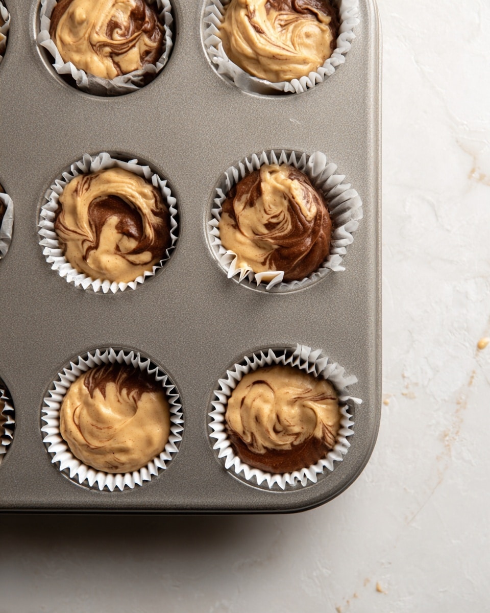 The image shows a close-up of a gray muffin tray holding nine cupcake liners filled with a two-tone batter that is swirled together. Each liner has a light tan and darker brown batter mixed in a marbled pattern with uneven swirls and spots, creating a textured look on the surface. The liners are white with ridged edges and fit snugly into the tray. The tray is placed on a white marbled textured surface visible at the corner. There are small drops and smudges of batter on the tray around the liners. Photo taken with an iphone --ar 4:5 --v 7