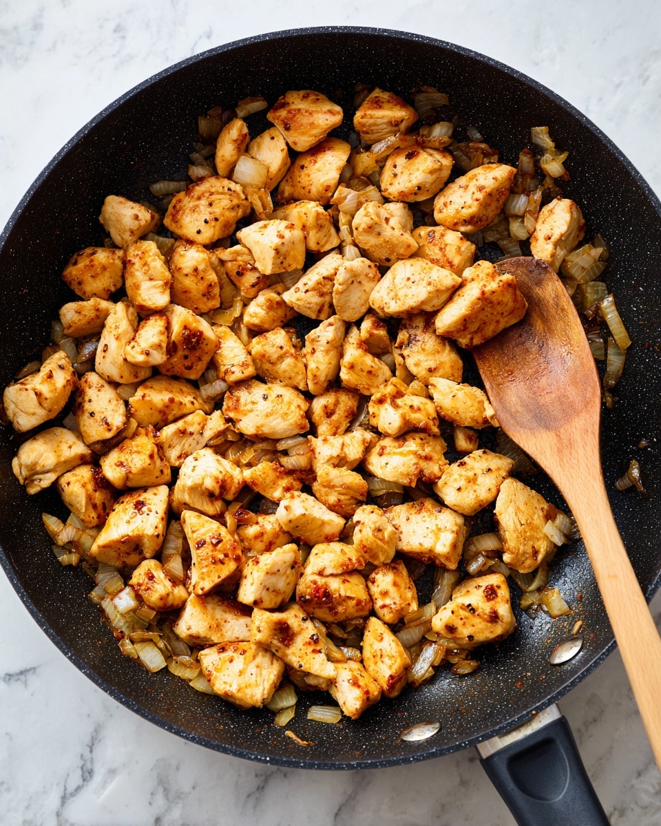 The image shows a black frying pan filled with many small, lightly browned pieces of cooked chicken. The chicken pieces are golden with some darker spice spots and mixed with small bits of sautéed onions that have a soft, translucent texture and a light brown color. A wooden spoon rests on the right side of the pan, partially under some chicken pieces. The background surface is a white marbled texture. photo taken with an iphone --ar 4:5 --v 7