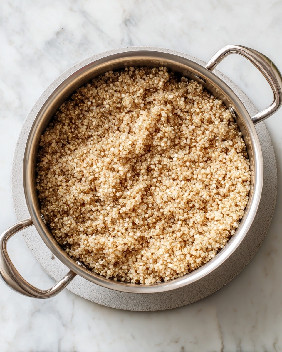 A top view of a stainless steel pot filled with cooked quinoa, small round grains showing a light brown color and fluffy texture, sitting on a light gray round trivet. The pot has two handles and is placed on a white marbled surface with subtle gray veins. photo taken with an iphone --ar 4:5 --v 7