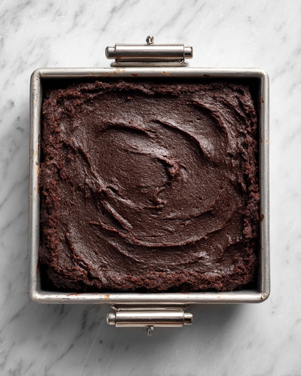A single thick layer of dark brown, almost black, dense brownie batter fills a square silver baking pan with four metal clips on the edges. The batter has a rough, slightly cracked surface texture with swirled patterns all over. The pan is set on a white marbled surface with subtle gray veining. photo taken with an iphone --ar 4:5 --v 7
