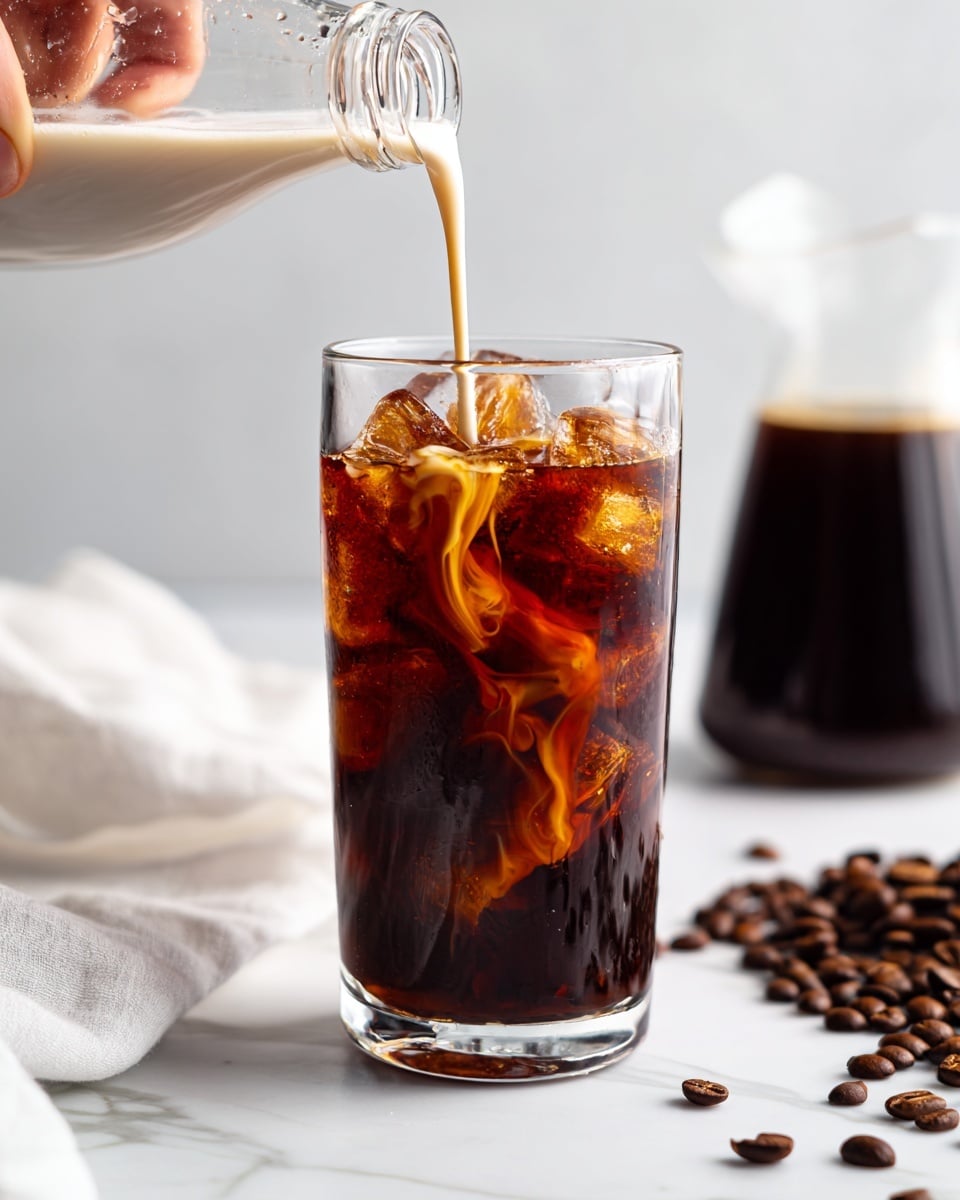 A clear glass filled with dark brown iced coffee and ice cubes sits on a white marbled surface, with a creamy white liquid being poured from a small glass bottle by a woman's hand, creating swirls and mixing with the coffee in the glass. In the background to the right, there is a French press filled with dark coffee. Scattered coffee beans lay on the white marbled surface in the lower right corner, and a white cloth is partially visible in the lower left. The overall setting is bright with a clean and minimal look. photo taken with an iphone --ar 4:5 --v 7