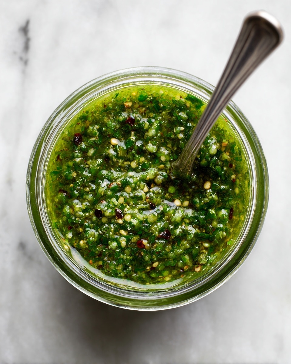 A close-up image of a clear glass jar filled with a textured green sauce that has small bits of herbs and spices visible, showing a mix of dark and bright green colors with hints of yellow and tiny red flakes. The sauce looks thick but not smooth, with an uneven surface that shows lumpiness and a fresh, herbal look. The jar sits on a gray cloth with frayed edges on a white marbled surface, with the focus on the top part of the jar and the sauce inside photo taken with an iphone --ar 4:5 --v 7