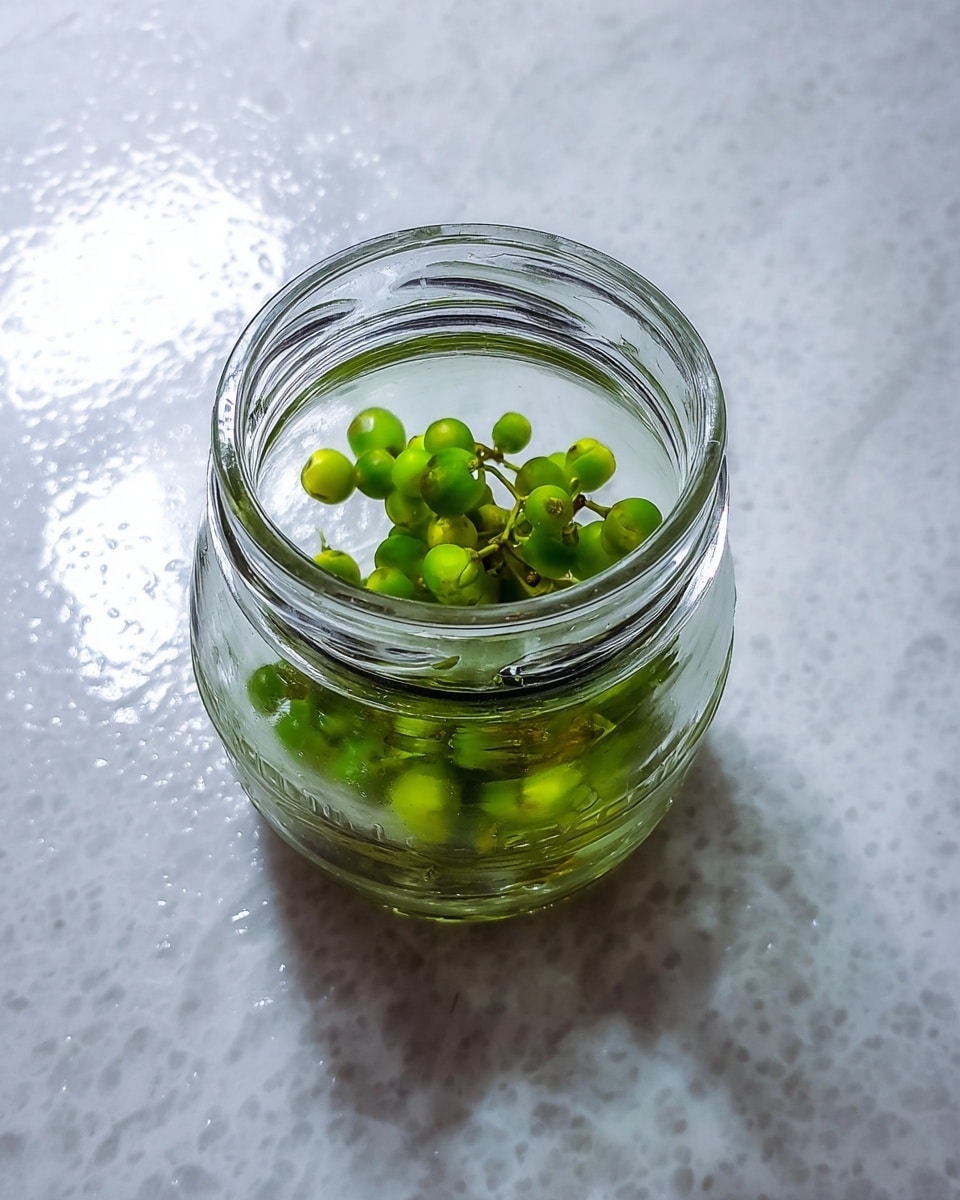 A clear small glass jar is shown sitting on a white marbled surface. Inside the jar, there are small green berry-like fruits or seeds clustered together near the bottom. The jar has an open mouth with visible thick glass rings at the top. The background surface has a smooth shiny texture with white specks, adding contrast to the transparent jar and green contents inside. photo taken with an iphone --ar 4:5 --v 7