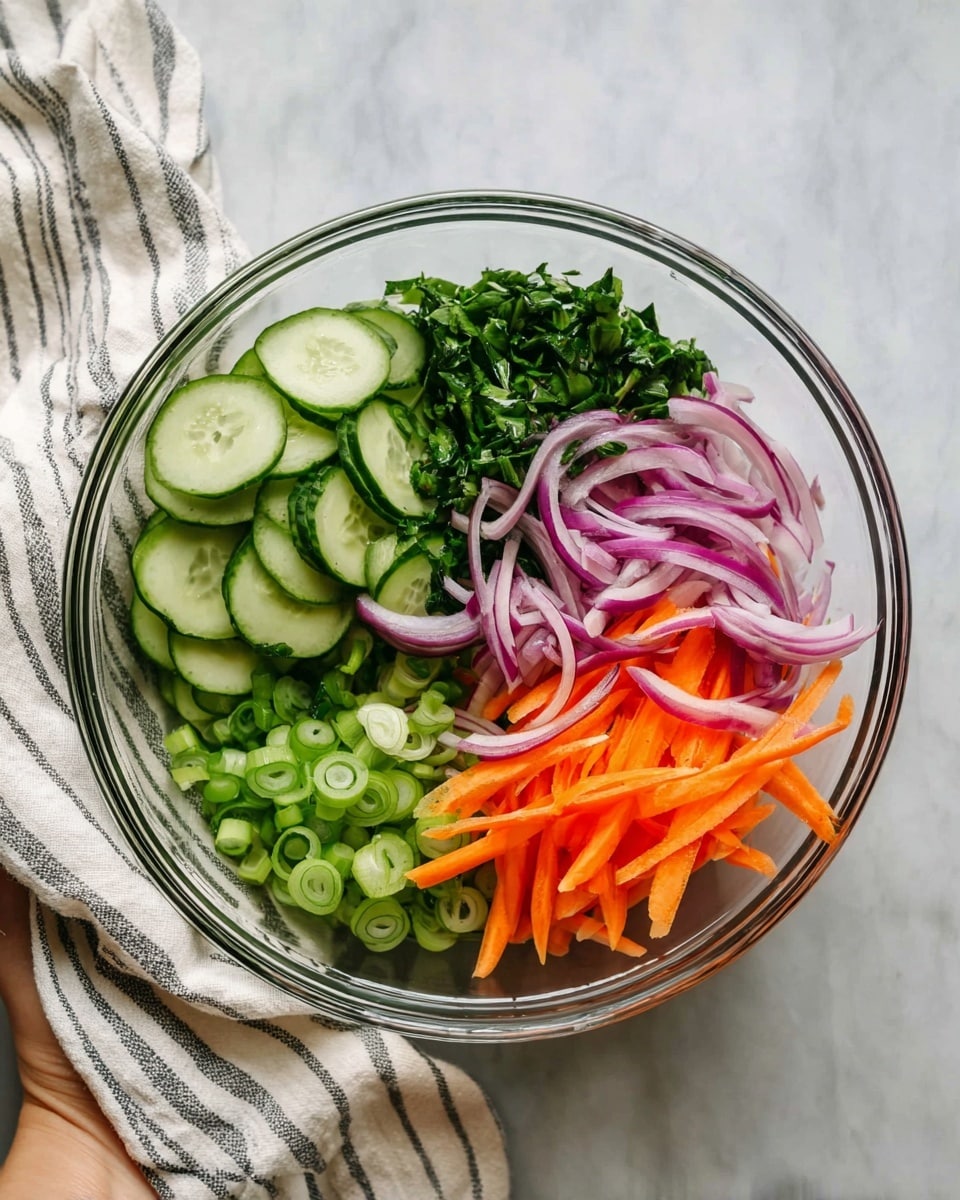 A clear glass bowl on a white marbled surface holds a fresh salad with five visible layers arranged side by side. On the left, there are thin cucumber slices, light green with darker green edges. Next to the cucumbers is a pile of chopped dark green leafy herbs. Above the herbs, there are thin, curved slices of purple-red onion. To the right of the onions, bright orange carrot sticks are stacked. In the center and spreading slightly into other sections, there are light green ring-shaped pieces of sliced green onion. A woman’s hand holds the bowl slightly from the left side, and a white cloth with black stripes rests nearby. Photo taken with an iphone --ar 4:5 --v 7