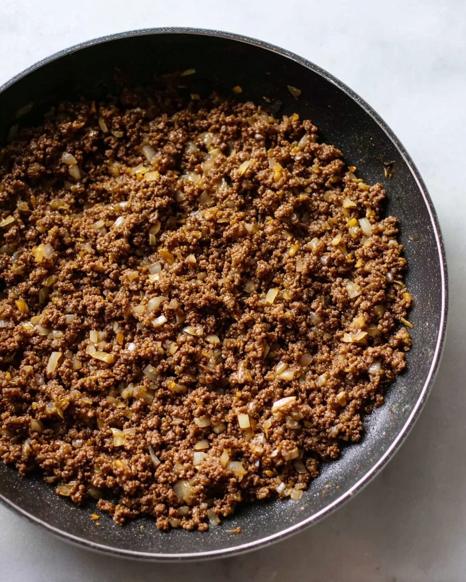 The image shows a close-up of cooked minced meat mixed with small pieces of cooked onion in a black pan. The meat is brown with varied textures, showing a crumbly and slightly moist appearance, while the onions add tiny yellowish and translucent bits scattered evenly through the mixture. The pan is round with a smooth black surface, sitting on a white marbled countertop. Photo taken with an iphone --ar 4:5 --v 7