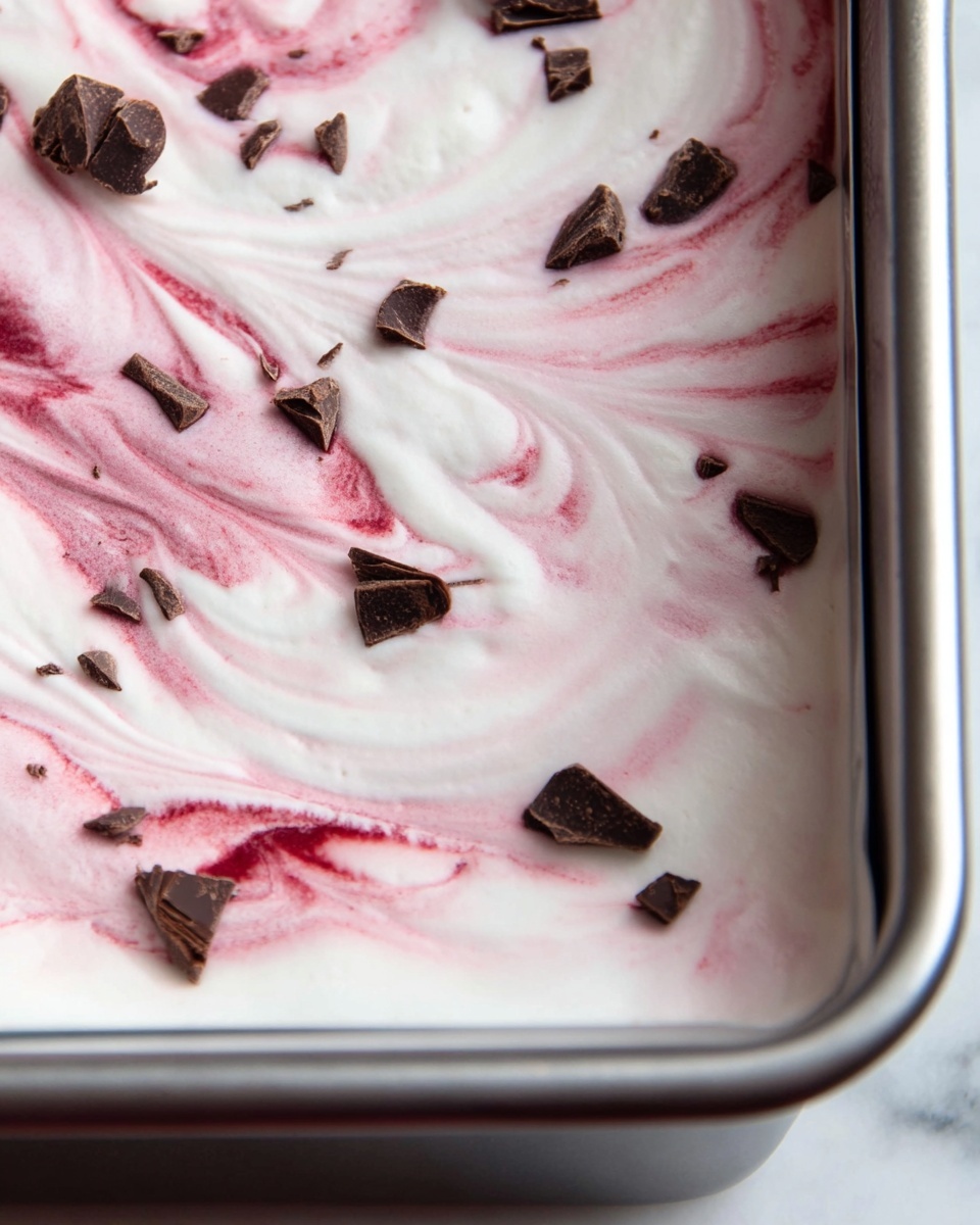 This image shows a close-up view of a dessert in a silver baking pan. The dessert has a smooth, white creamy layer spread evenly across the surface. Swirled through the white cream are soft pink and red streaks, creating a marbled effect. Scattered on top are small, dark brown chocolate pieces that add texture and contrast to the creamy white and pink layers. The background features a white marbled surface beneath the pan. photo taken with an iphone --ar 4:5 --v 7