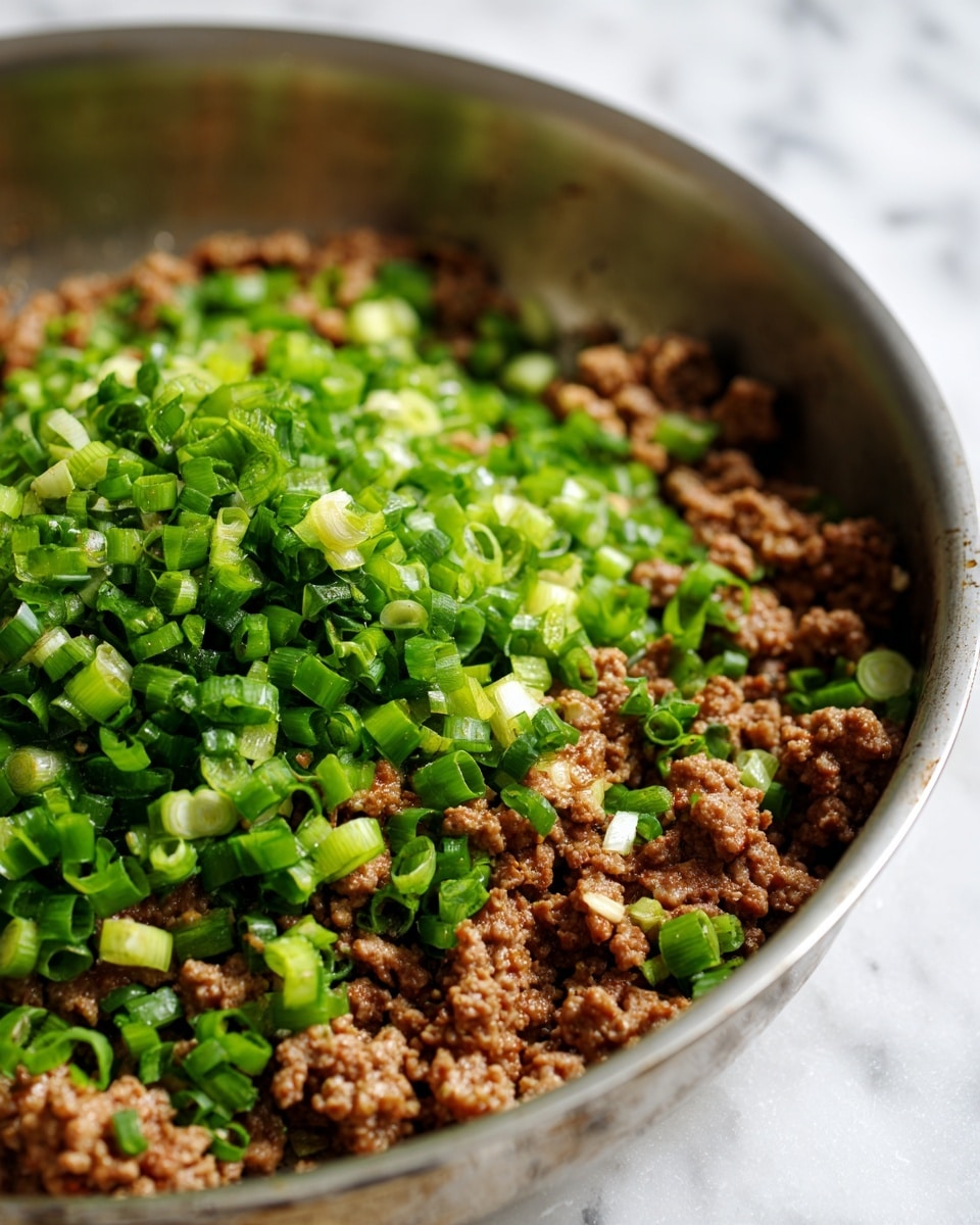 The image shows a close-up of cooked ground meat with a rough texture in a pan. On top, there is a thick layer of chopped green onions with a mix of bright and light green colors, adding freshness and contrast to the brown meat underneath. The meat appears moist with some small visible bits of fat and juices spreading slightly at the edges. The pan has a shiny metal surface with a soft focus background of a white marbled texture. Photo taken with an iphone --ar 4:5 --v 7