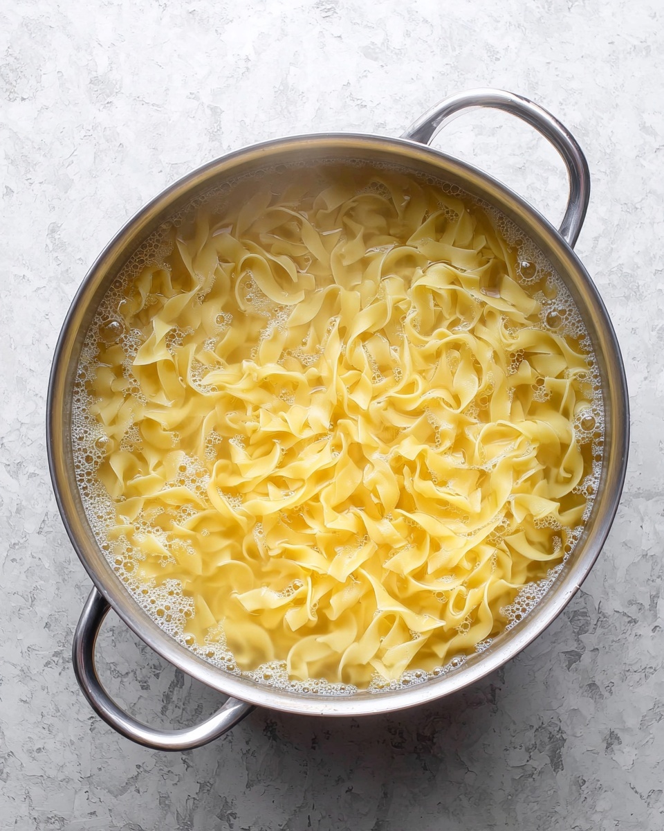 A silver metal colander filled with cooked yellow egg noodles that are thick and curly, with steam rising from the pasta, sitting on a white marbled surface texture. The colander has small round holes and two handles on each side, showing the shiny inside metal. photo taken with an iphone --ar 4:5 --v 7
