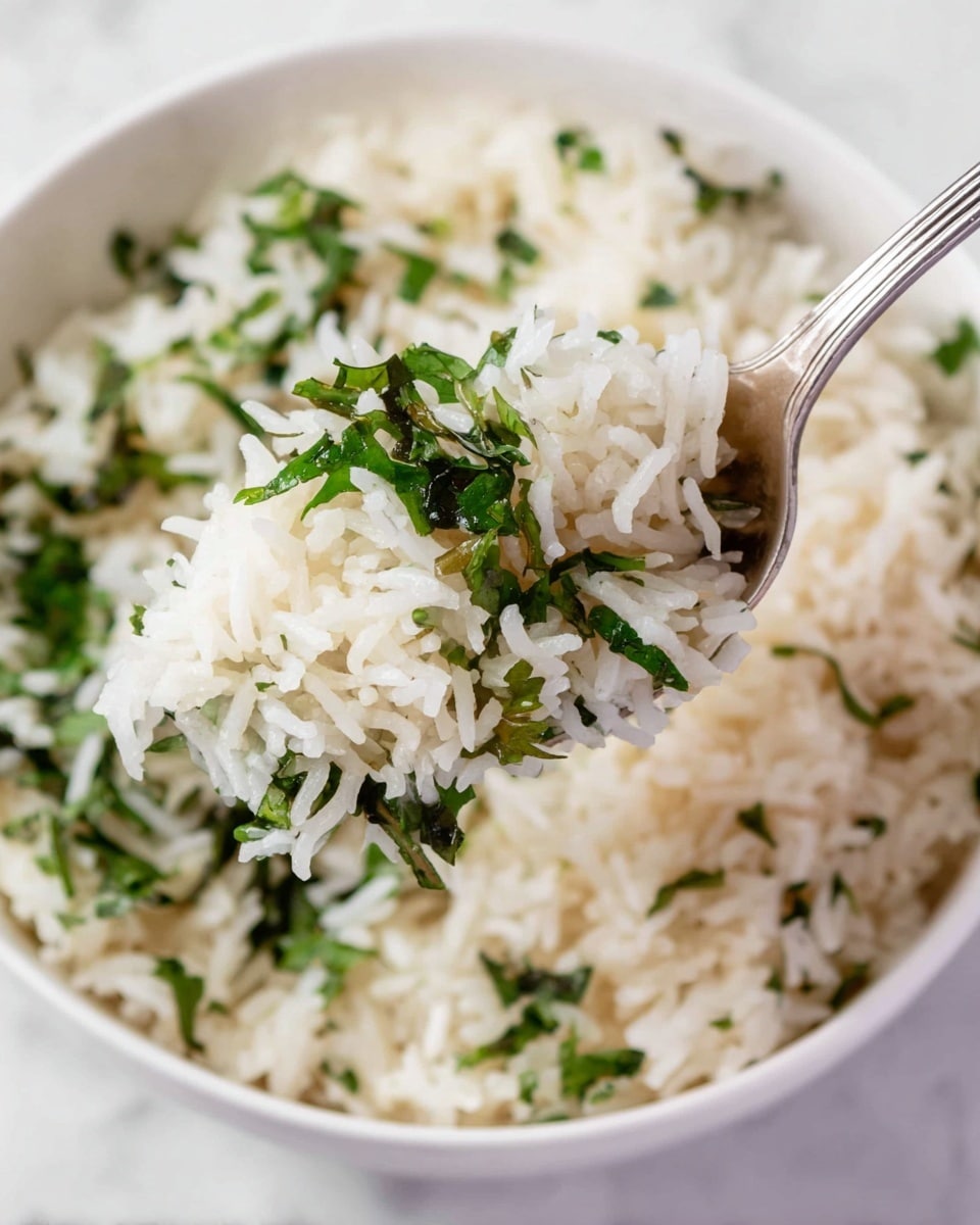 A close-up view of a white bowl filled with two main layers: the base layer is a heap of fluffy white rice with long grains, and the top layer shows scattered pieces of green leafy herbs mixed into the rice. A silver spoon holds a portion of the rice and herbs above the bowl, highlighting the texture of the soft grains and fresh green leaves. The scene is set on a white marbled surface. photo taken with an iphone --ar 4:5 --v 7