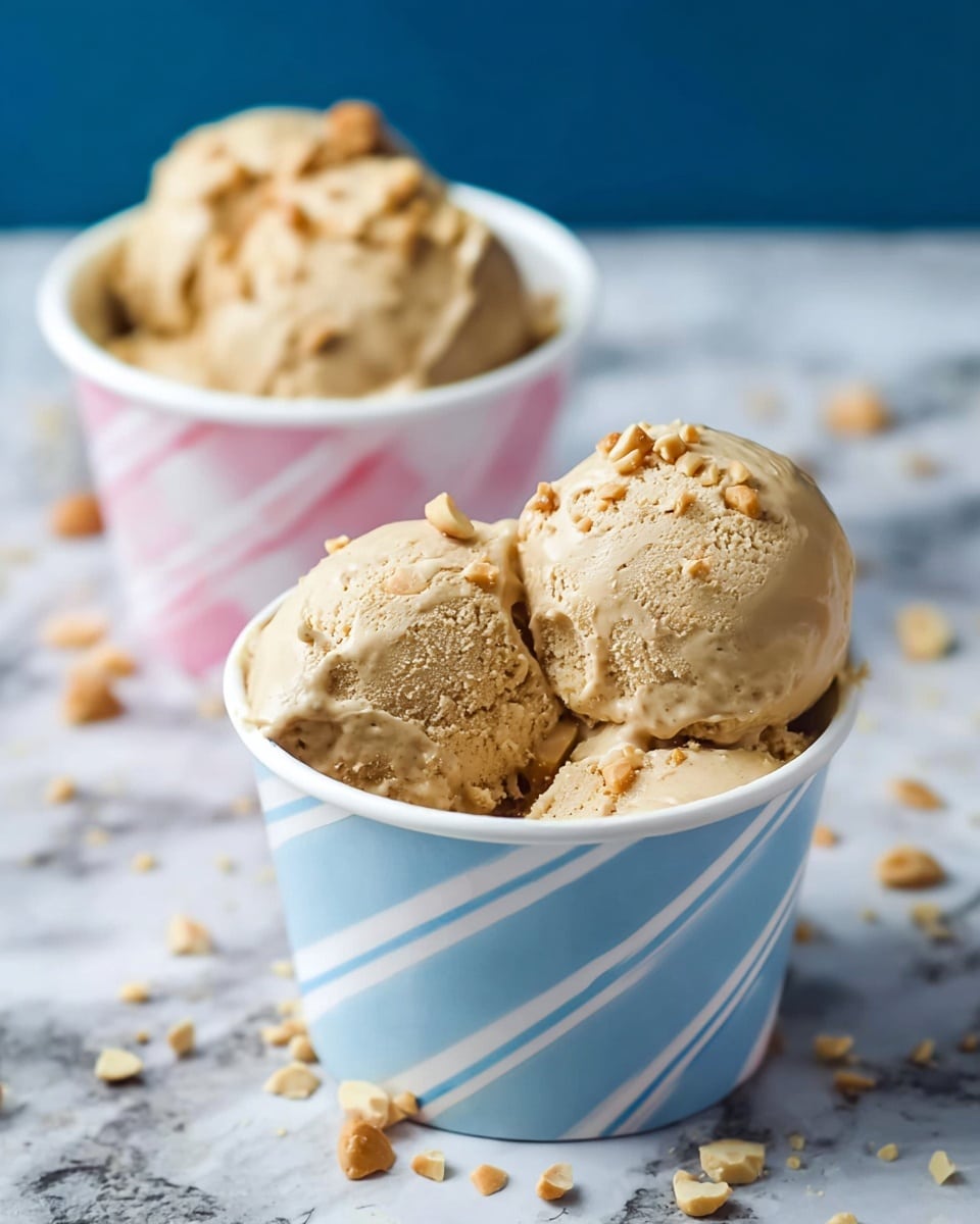 Two cups of ice cream are shown, each with three scoops of tan-colored ice cream with small nut pieces mixed in. The ice cream has a creamy, slightly rough texture. The cup in the front is white with blue diagonal stripes and is filled with the ice cream scoops and sprinkled nuts on top. Behind it, another cup with pink and white diagonal stripes is partially visible, also filled with the same ice cream. The cups are placed on a white marbled surface with scattered small nut pieces around. Photo taken with an iphone --ar 4:5 --v 7