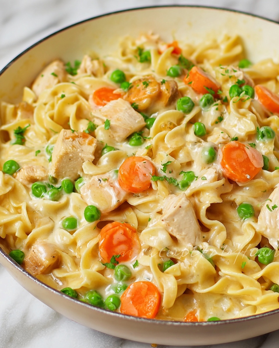 This image shows a close-up of a creamy noodle dish in a white pan placed on a white marbled surface. The dish has three main layers: the base layer consists of soft, twisted egg noodles in a light cream sauce; scattered throughout are pieces of cooked, light brown chicken chunks adding texture and color contrast; on top, there are bright orange carrot slices and fresh green peas spread evenly, adding vibrant pops of color. The creamy sauce gives a smooth texture coating the noodles and vegetables. The overall look is warm and inviting with mixed textures and fresh colors. photo taken with an iphone --ar 4:5 --v 7