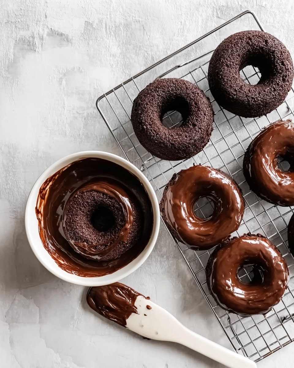 The image shows a baking scene with five chocolate doughnuts arranged on a cooling rack over a white marbled surface. Three doughnuts are plain with a dark, rough texture, and two are coated in a shiny, smooth chocolate glaze. One doughnut is placed inside a white bowl that contains thick chocolate glaze. A white spatula with some chocolate is lying next to the bowl on the white marbled surface. The overall look is calm and focused on the rich chocolate doughnuts. photo taken with an iphone --ar 4:5 --v 7