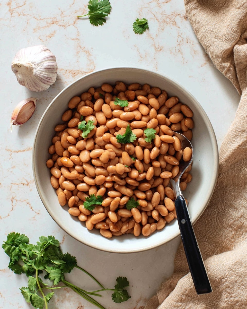 A white bowl filled with a large pile of smooth, light brown beans that have a slightly shiny surface, with a few small green cilantro leaves scattered on top for color contrast. A spoon with a black handle rests inside the bowl on the right side, partially submerged in the beans. The bowl sits on a white marbled surface, and some garlic cloves and a light beige cloth napkin are visible around the bowl, adding natural textures and a rustic feel to the scene. Photo taken with an iphone --ar 4:5 --v 7