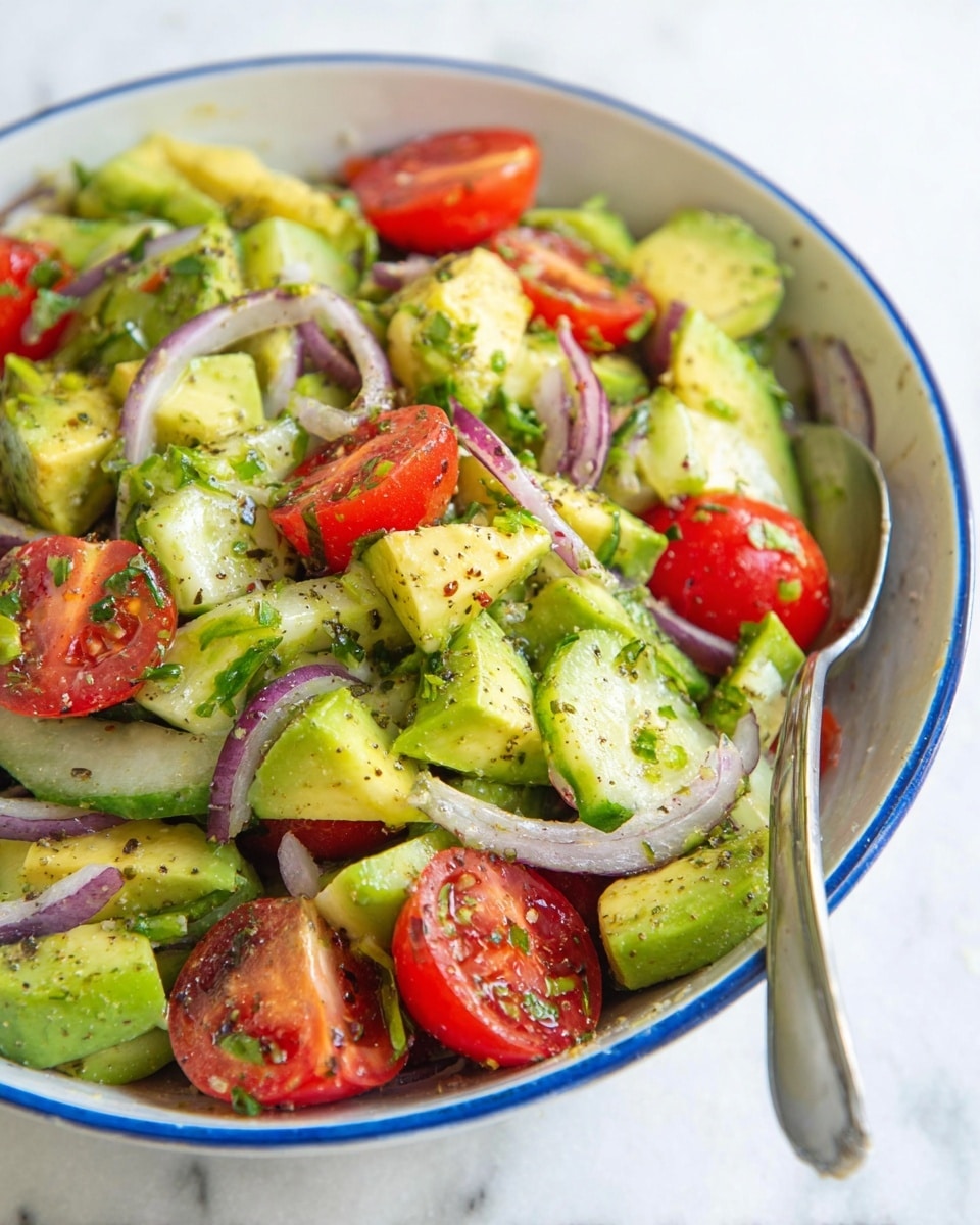 A close-up of a mixed salad placed in a white bowl with a blue rim, showing layers of green avocado chunks, bright red cherry tomato halves, white cucumber pieces, thin slices of purple onion, and chopped green onions, all mixed and lightly seasoned with pepper and herbs. A silver spoon is visible on the right side of the bowl. The bowl sits on a white marbled surface. Photo taken with an iphone --ar 4:5 --v 7