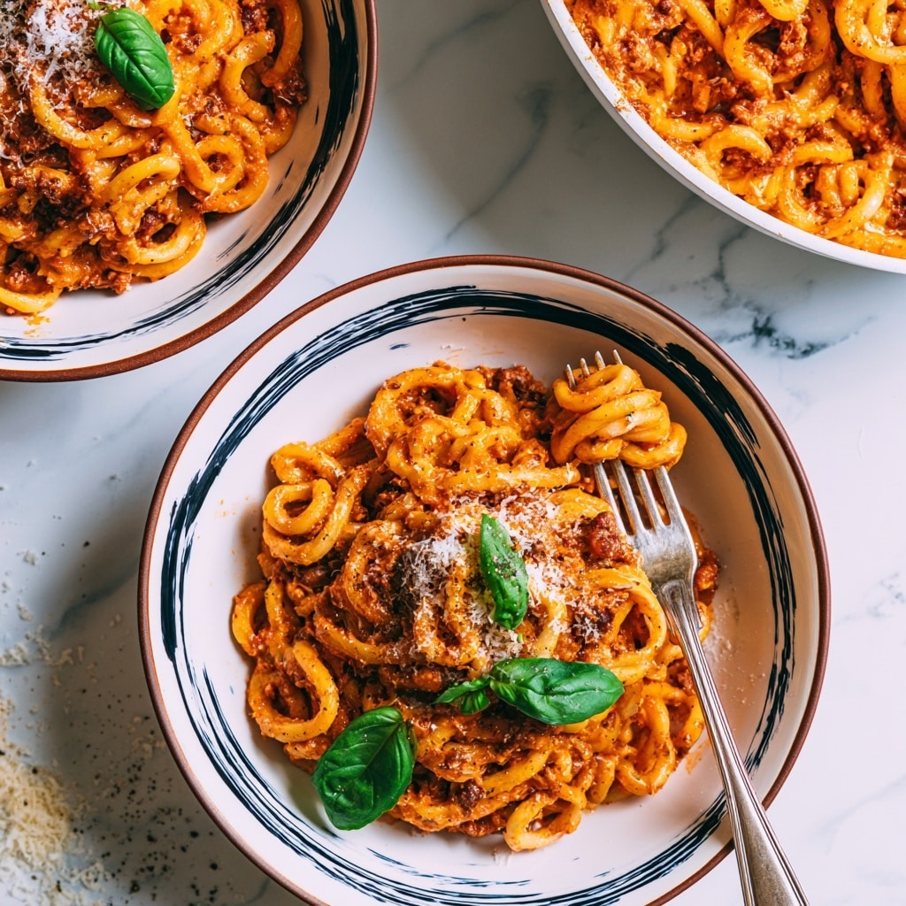 Two white bowls with dark blue brushstroke patterns and a brown rim hold a creamy orange pasta with meat sauce and grated cheese on top. The pasta is thick and twisted, covered evenly in sauce, with bits of meat visible in the mix. Each bowl is placed on a white marbled surface, with bright green basil leaves on the pasta adding color contrast. A silver fork is stuck in one bowl, showing some pasta twirled around it. On the top right, a white pan holds more of the same creamy pasta with meat sauce. The whole scene is bright and inviting with scattered cheese and black pepper nearby, photo taken with an iphone --ar 4:5 --v 7