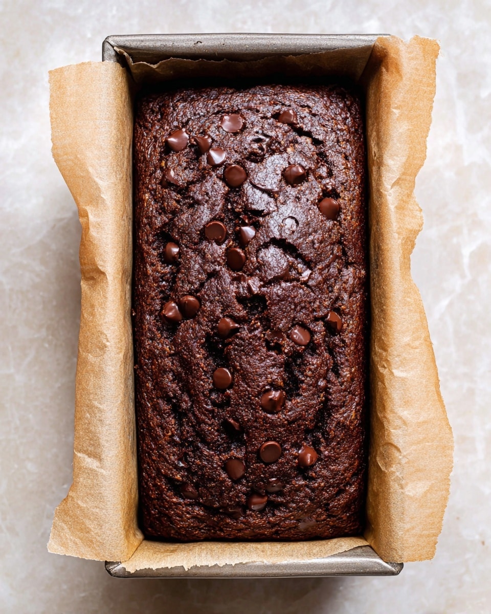 A close-up top view of a dark brown chocolate loaf cake in a rectangular metal pan lined with light brown parchment paper. The cake surface is textured with small cracks and scattered dark chocolate chips on top, adding a slight glossy look. The pan sits on a white marbled surface. photo taken with an iphone --ar 4:5 --v 7