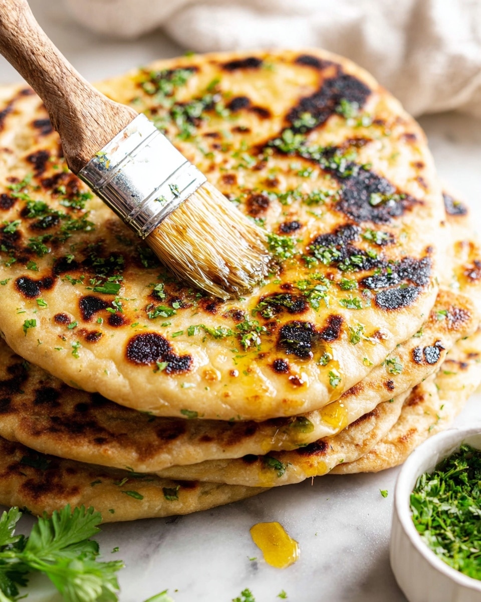 A stack of five golden brown flatbreads with dark char marks is placed on a white marbled surface, with bright green chopped herbs sprinkled on top and between layers. The top flatbread is being brushed with a shiny golden oil using a large brush with a wooden handle, making the surface glisten. In the background, a white bowl filled with more green herbs is partially visible on the right side, and some fresh parsley leaves add color near the bottom left corner. The textures of the flatbreads show soft, slightly puffy areas with rougher browned spots. photo taken with an iphone --ar 4:5 --v 7