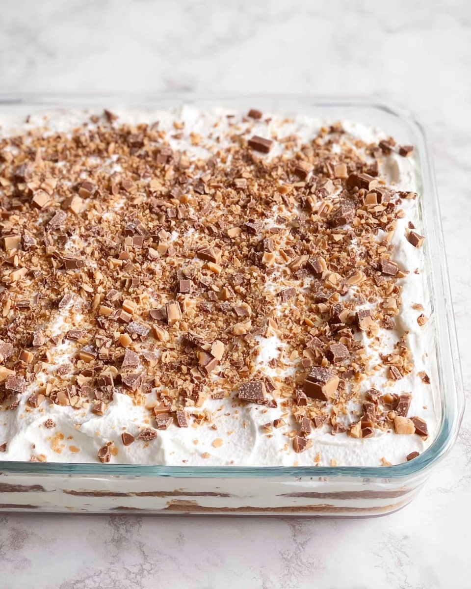 A glass rectangular dish filled with a layered dessert. The bottom layer is creamy white, covered by a thick white layer that looks like whipped cream or frosting. The top layer is sprinkled evenly with small chunks of light and dark brown pieces, which seem like chopped candy or nuts scattered all over. The dish is placed on a white marbled surface. photo taken with an iphone --ar 4:5 --v 7