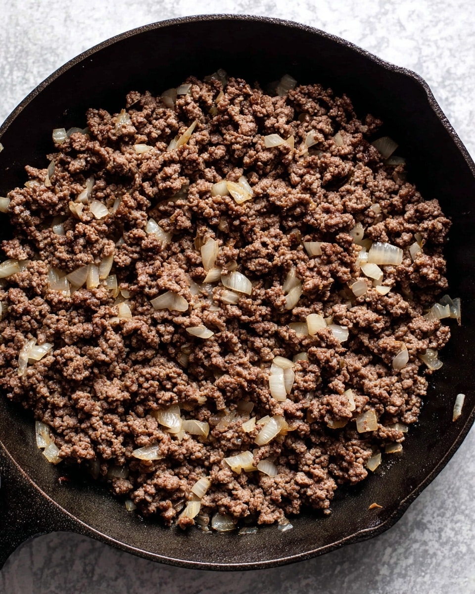 A close-up top view of cooked ground meat mixed with soft, translucent small chopped onions inside a dark cast iron skillet, the meat is brown and crumbly with some pieces showing slight texture from the onions, the skillet rests on a white marbled textured surface, the dark skillet contrasts with the lighter background photo taken with an iphone --ar 4:5 --v 7