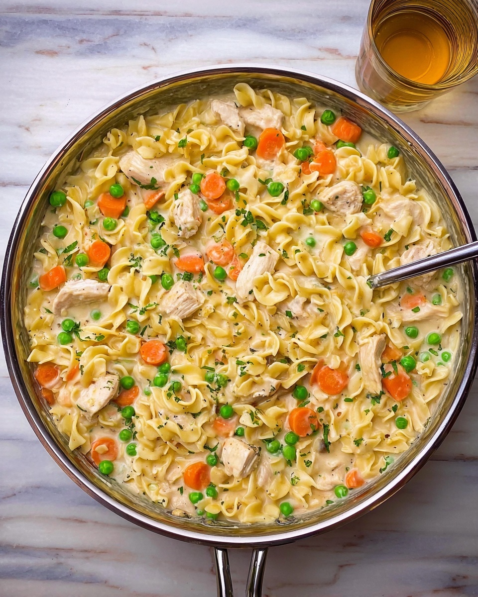 A large pan filled with creamy chicken and noodles sits on a white marbled surface. The dish has three main layers visible: at the base, there is a creamy white sauce that looks thick and smooth. Spread throughout are light yellow egg noodles that are soft and slightly curled. Mixed evenly in are small chunks of cooked chicken with a light golden-brown color, bright orange slices of cooked carrot, and scattered bright green peas, adding color and texture to the dish. The pan has a shiny metal rim and handle, and the food inside looks warm and comforting. photo taken with an iphone --ar 4:5 --v 7