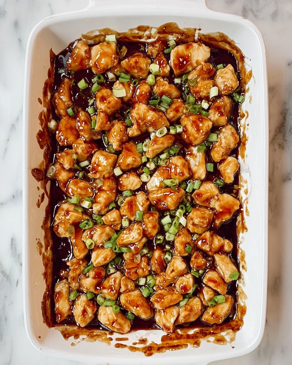 A white rectangular baking dish filled with many small pieces of light golden chicken coated in a thick shiny dark brown sauce, scattered with small pieces of bright green chopped scallions on top; the sauce smudges the sides of the dish with some splashes visible around the top edge; the dish sits on a white marbled surface photo taken with an iphone --ar 4:5 --v 7