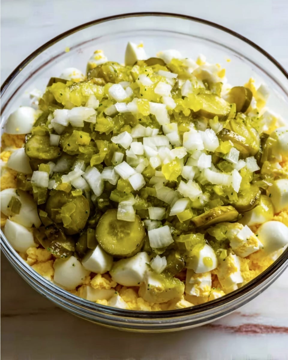 A clear glass bowl filled with three layers of ingredients is shown on a white marbled surface. The bottom layer consists of small yellow and white pieces, likely chopped boiled eggs. The middle layer is made up of light green slices of pickles spread evenly over the eggs. The top layer is a mix of finely chopped white onions and diced pickles, giving a fresh and crunchy texture to the dish. Photo taken with an iphone --ar 4:5 --v 7