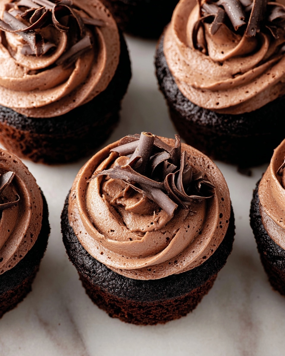 A close-up view of several chocolate cupcakes arranged on a white marbled surface, each cupcake having two layers with a dark, moist base and a thick, smooth, light brown chocolate frosting layer swirled on top. The frosting has a soft, creamy texture with tiny air bubbles visible, and each cupcake is topped with delicate, curled chocolate shavings that add a decorative and rich finishing touch. The cupcakes fill most of the frame, showing the fine texture and chocolate details clearly. photo taken with an iphone --ar 4:5 --v 7