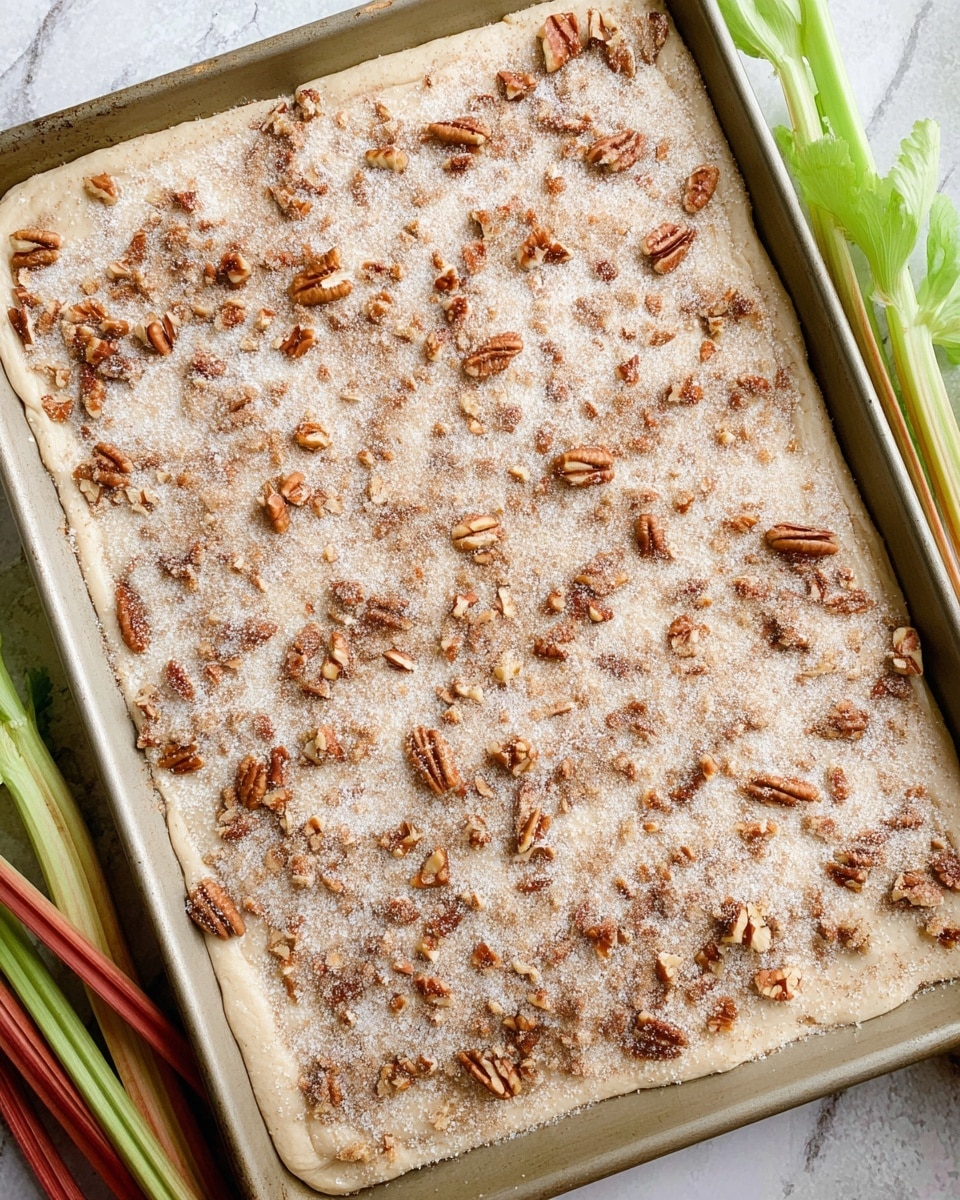 The image shows a metal rectangular pan filled with a thick, creamy batter that has small pieces of light green and red mixed in, spread evenly to cover the surface. Behind the pan, there are fresh stalks and leaves of rhubarb with green leaves and red stems, resting on a white marbled surface. The batter texture looks smooth with some swirls from spreading, and the pan is placed at an angle, showing part of the mixture close up. photo taken with an iphone --ar 4:5 --v 7