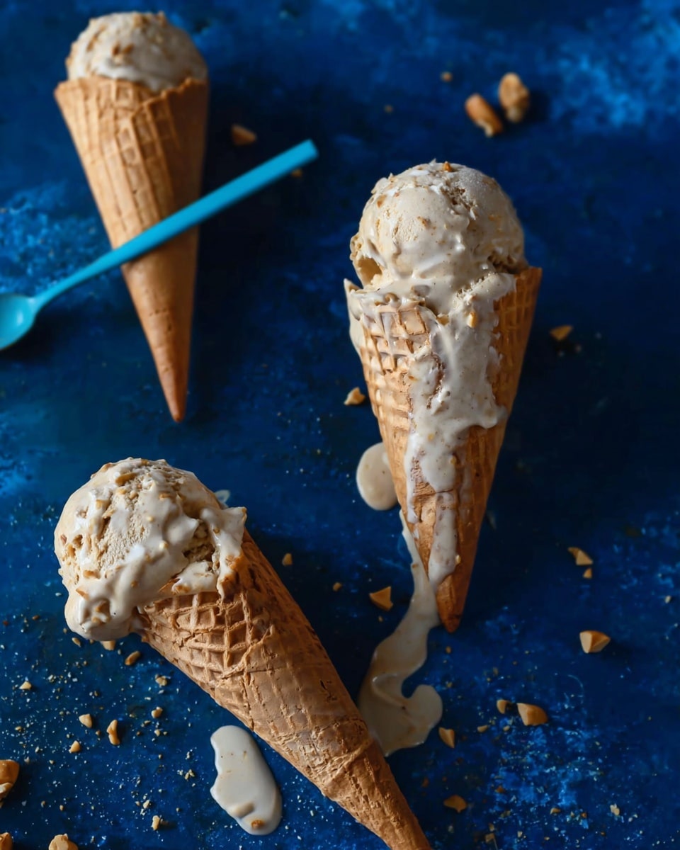 Three waffle cones are filled with melting light brown ice cream with small nut pieces mixed throughout. One cone is upright with a scoop on top dripping slightly over the side, while the other two lie on their sides showing melting ice cream dripping onto a dark blue textured surface. Small nut pieces are scattered around the cones, and a thin light blue spoon is visible on the left side. The background has a dark blue, soft texture. photo taken with an iphone --ar 4:5 --v 7