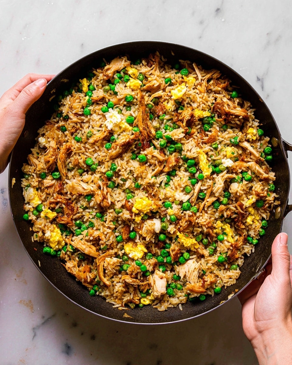 A large black pan is filled with a colorful mix of fried rice layered with shredded cooked chicken and bright green peas scattered evenly throughout. There are small, fluffy yellow egg pieces mixed in, adding more color and texture. The chicken pieces show some browning, giving a rich contrast to the light brown rice. The pan is held by a woman's hand on each side, over a white marbled surface. The scene is brightly lit to highlight the different ingredients and textures. photo taken with an iphone --ar 4:5 --v 7