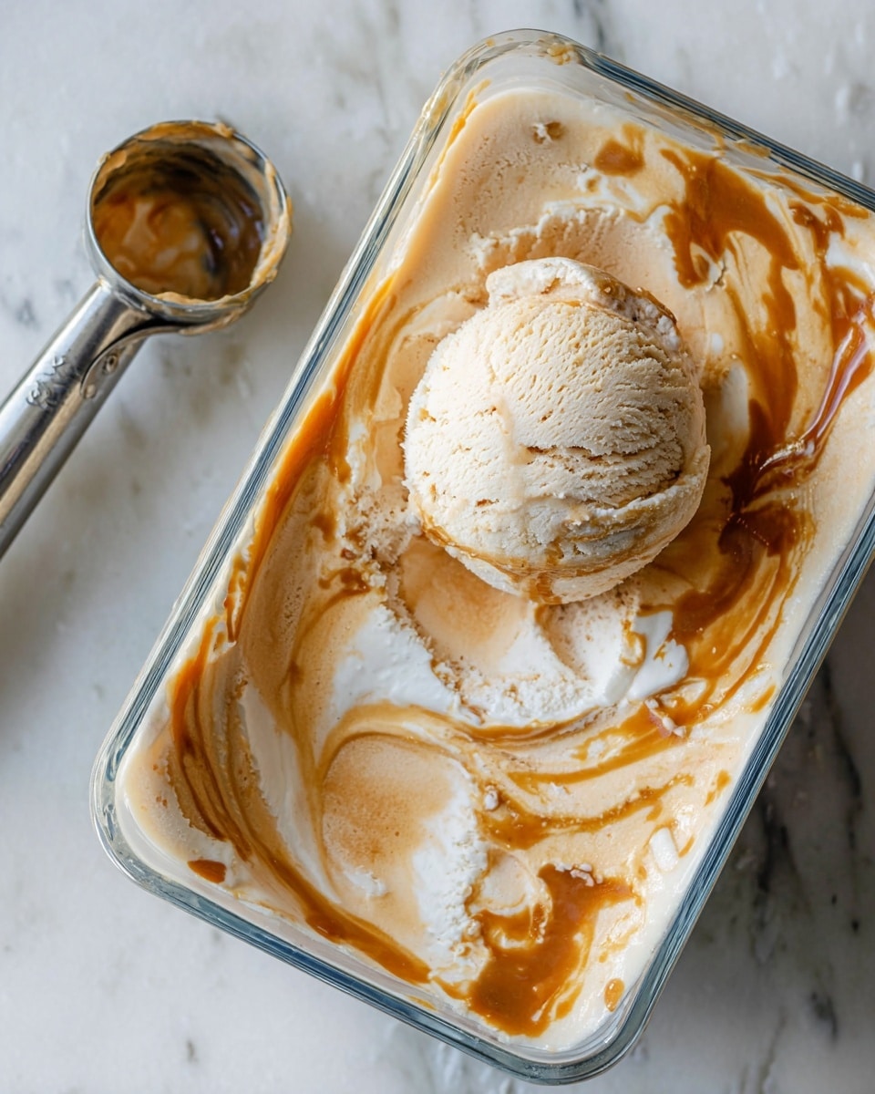 The image shows a glass container filled with creamy ice cream that has two main layers swirled together: a light beige layer and a caramel brown layer, creating a marbled effect. A round scoop of the ice cream rests on top, displaying a slightly textured and soft surface from being scooped out. The container sits on a white marbled surface. Near the container, there is an ice cream scooper with traces of the ice cream inside the metal scoop, its handle resting on the surface. Photo taken with an iphone --ar 4:5 --v 7