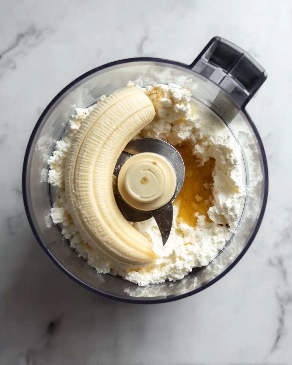 A clear food processor bowl shows a peeled banana lying on top of a layer of white cottage cheese, with a small amount of golden honey drizzled beside the cheese. The bowl is placed on a white marbled surface, and the blade of the processor is visible in the center, dividing the ingredients. The scene is bright and clean, highlighting the textures of the soft banana skin, crumbly cottage cheese, and shiny honey. Photo taken with an iphone --ar 4:5 --v 7