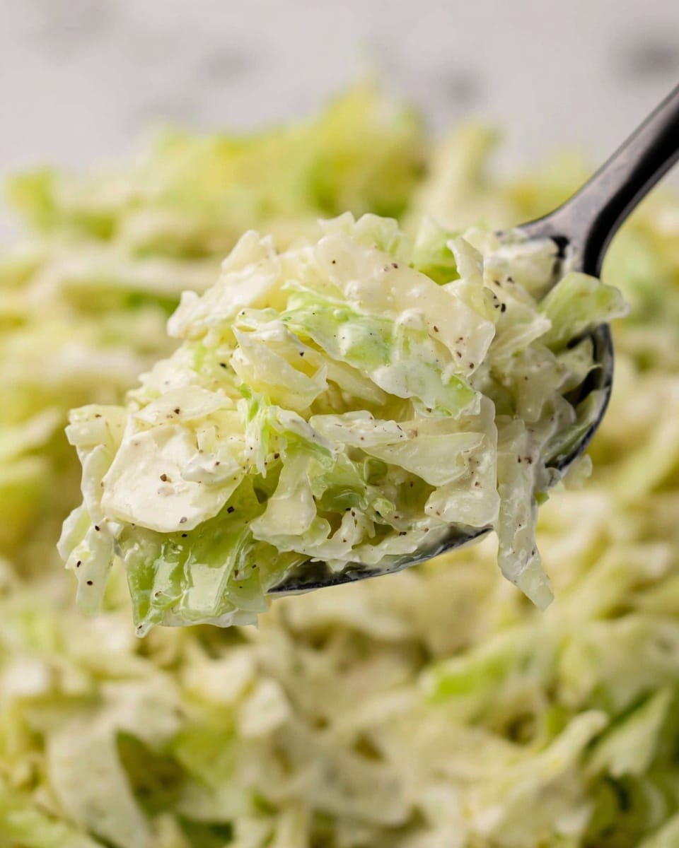 A close-up view of a spoon filled with a mix of finely shredded cabbage and a creamy white dressing, showing small black pepper pieces sprinkled throughout. The mix is layered with light green and white cabbage shreds that have a slightly wet texture from the dressing. In the background, there is more of the same cabbage mix softly blurred to keep the focus on the spoon. The overall colors are pale green, white, and light cream, with small dark spots of pepper. The surface beneath is a white marbled texture. photo taken with an iphone --ar 4:5 --v 7