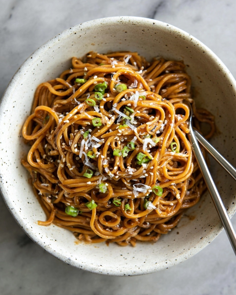 A bowl filled with a mound of cooked noodles that are light brown and glossy from sauce, topped with finely grated white cheese and small bits of green onion scattered over the top. The noodles are thick and twisted together tightly in layers, showing some texture and shine. The bowl is white with small brown spots and placed on a white marbled surface. Part of a cloth with stripes is blurry in the background, and part of a metal fork is inside the bowl on the right side. Photo taken with an iphone --ar 4:5 --v 7