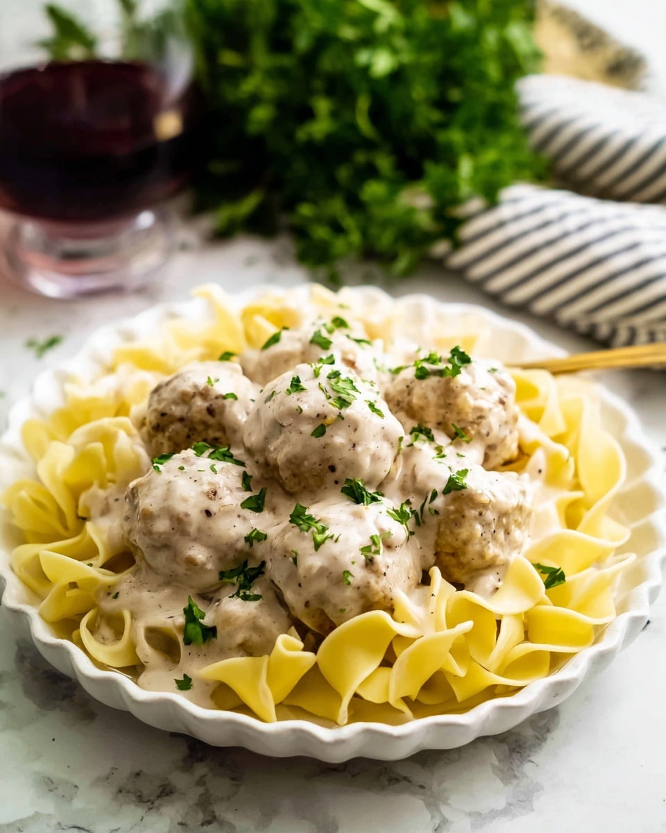 A silver pan filled with round, light brown meatballs covered in a creamy, pale beige sauce, sprinkled with small pieces of bright green herbs. Above the pan is a clear glass bowl full of plain cooked pasta with a light yellow color and smooth, slightly curled noodles. On the left side, there is a bunch of fresh green parsley resting on a white cloth with thin black stripes, all placed on a white marbled surface. Some green herb bits are scattered around the pan. photo taken with an iphone --ar 4:5 --v 7