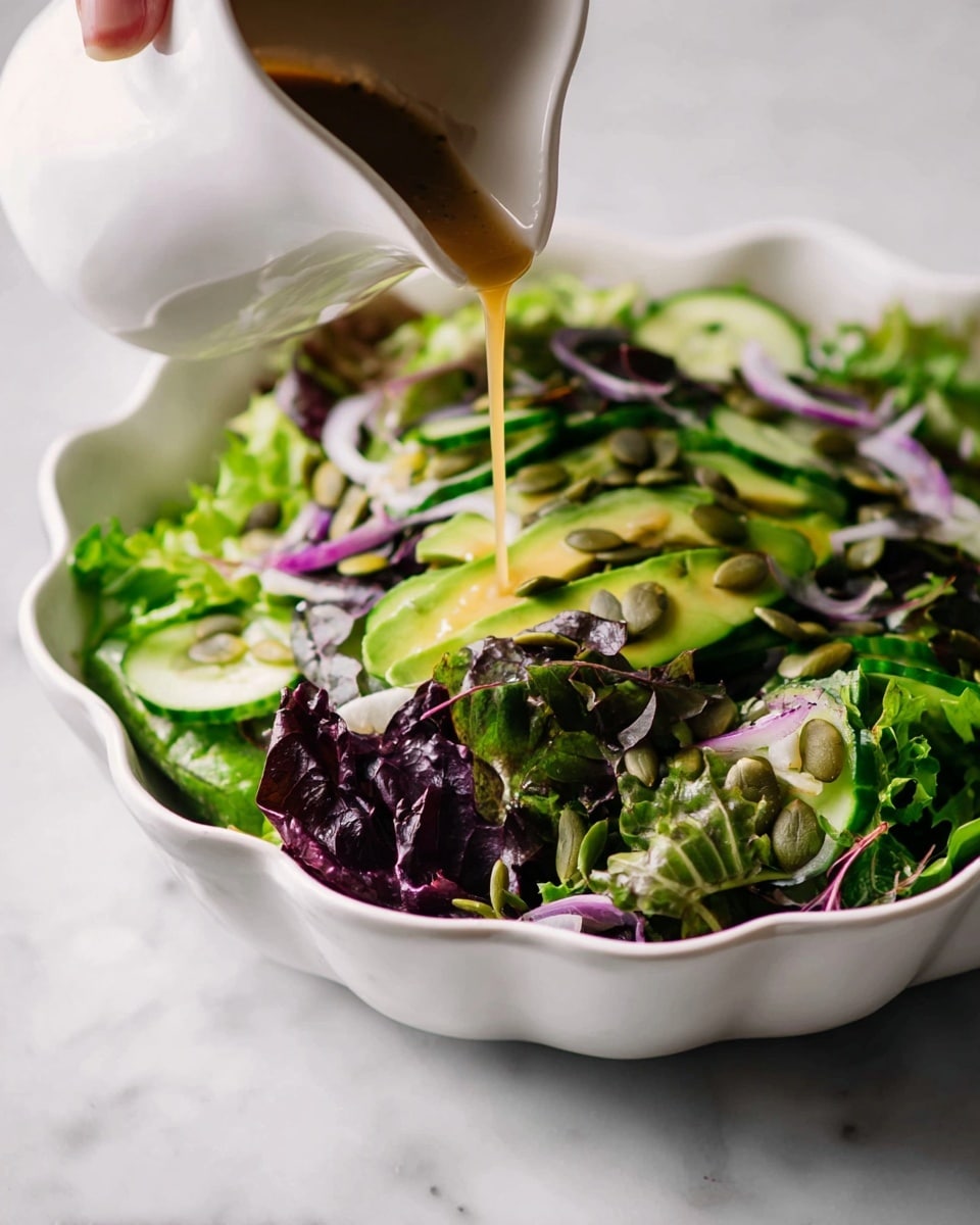 A white scalloped bowl holds a fresh mixed green salad with three main layers: the base layer is a mix of leafy greens with deep green and purple hues, the middle layer includes bright green cucumber slices and purple onion slivers, and the top layer is sprinkled with light brown pumpkin seeds. A light brown dressing is being poured over the salad from a white pitcher, with a woman's hand steadying it. The scene is set on a white marbled surface. photo taken with an iphone --ar 4:5 --v 7