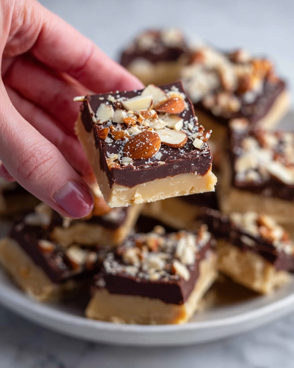 A woman's hand holds a square piece of a layered dessert, showing three clear layers: a light tan base with a smooth texture, a thick dark brown chocolate layer in the middle, and a topping of chopped almonds scattered unevenly on the glossy chocolate surface. The rest of the square pieces with the same layers are piled in a white bowl on a white marbled surface in the background. Photo taken with an iphone --ar 4:5 --v 7