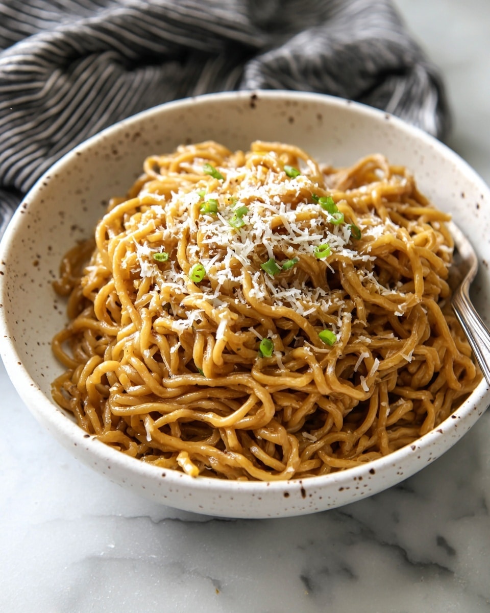 A white speckled bowl filled with a large pile of brown noodles coated in sauce. The noodles have a shiny, smooth texture and are topped with small white sesame seeds, light green chopped scallions, and fine white shavings. Two metal chopsticks rest in the bowl on the upper right side. The bowl is placed on a white marbled surface. photo taken with an iphone --ar 4:5 --v 7