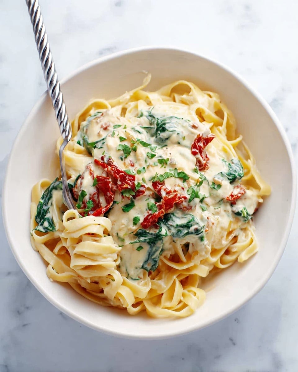 A white bowl sits on a white marbled surface, filled with three layers: the bottom layer is a nest of light yellow fettuccine pasta with a smooth texture, next comes a creamy white sauce with visible green spinach leaves and red sun-dried tomato strips stirred in, creating a soft and slightly chunky texture, and the top layer is a sprinkling of finely chopped green herbs. A twisted silver fork is placed inside the pasta on the left side, slightly twirling the noodles. Photo taken with an iphone --ar 4:5 --v 7